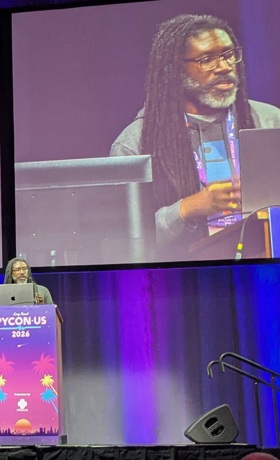 Lawrence Gray speaking at a lectern at PyCon US 2026, with his face shown on the large screen behind him